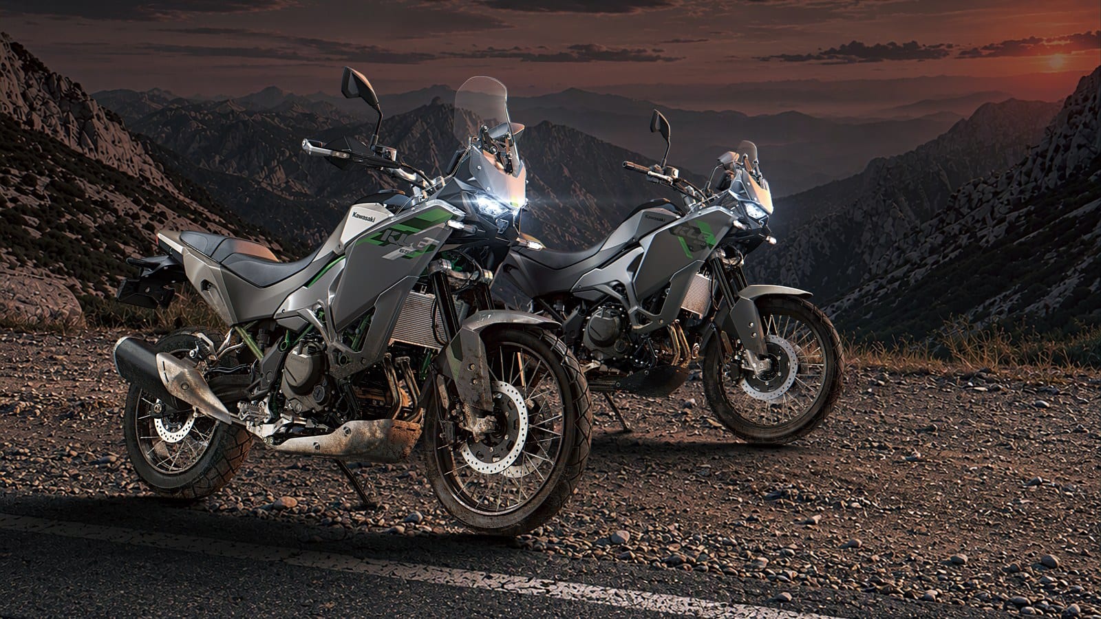 Two gray adventure motorcycles with green accents, spoked wheels and tall windscreens parked on gravel beside a paved road, with mountains and a sunset sky behind.