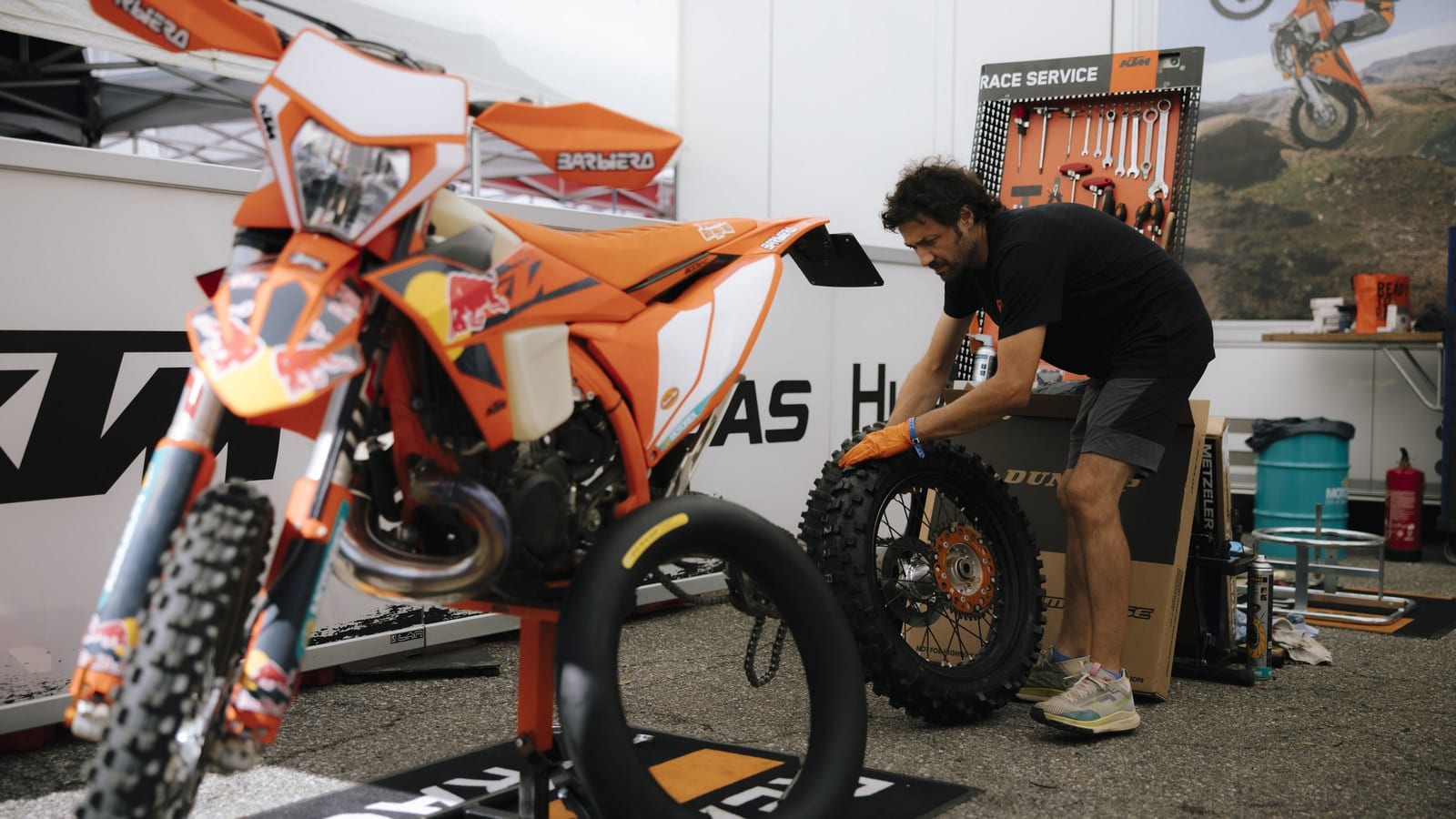 An orange KTM enduro bike with its rear wheel removed sits on a stand in a KTM service area while a mechanic handles the knobby rear wheel beside a Race Service tool board
